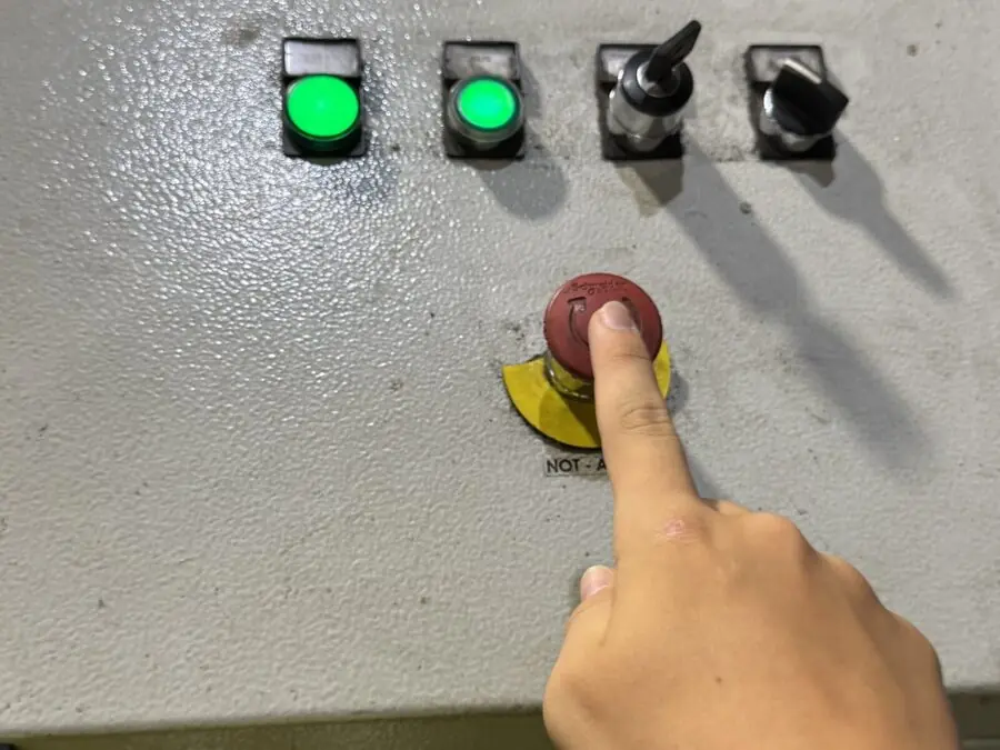 Eine Hand am Bedienpult der Anlage, der Finger liegt auf dem Not-Aus neben zwei grün leuchtenden Tastern